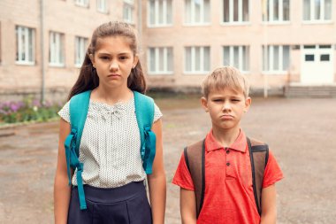 İki mutsuz öğrenci, farklı yaşlarda öğrencileri üzüyor. Okulun önünde dikilip kameraya bakıyorlar.