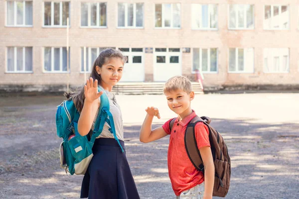 Little boy and his elder sister waving goodbye to parents on their way to school, standing in front of building of conventional school