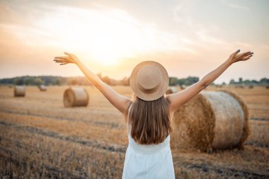 Beyaz elbiseli ve hasır şapkalı genç bir kız ellerini güneşe kaldırıyor, hasat edilen büyük bir tarlada saman balyalarıyla duruyor. Özgürlüğün sembolü, doğa, uyum, mutluluk.