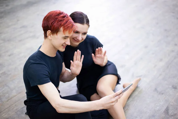 Two friends dancers in black clothes sitting on the floor and waving to other friend, while having a video chat. Virtual communication, modern technologies.