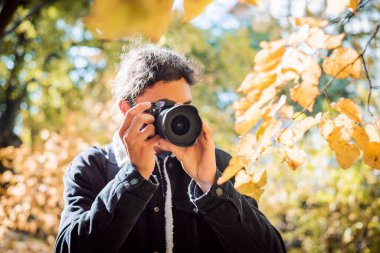 Young man photographer with dark curly hair in autumn park. Gorgeous sunny weather, bright lightened plants surround man.