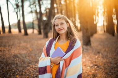 Young beautiful girl with a violet book of short stories in Fall park. Cheerful girl having a walk in park full of vibrant autumn colours, reading a book, admiring beauty of nature