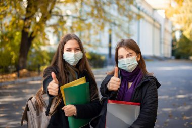 Two female students in protective medical masks showing thumbs up to the camera standing in university campus. Education during Covid-19 pandemic