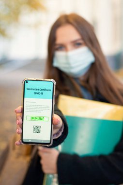 Beautiful student girl in medical mask showing electronic version of vaccination certificate to the camera. Vaccination among students and teachers of university concept