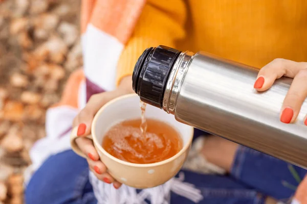 Person sitting outdoors and pouring tea from the thermos into white cup. Chilly autumn weather, warm drink in a walk on nature.