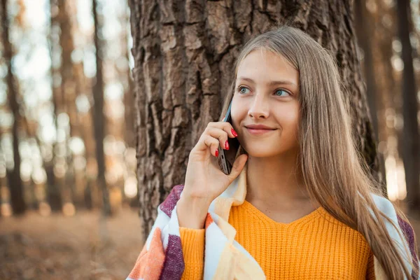 Cheerful teenage blond girl wearing orange sweater, covered in bright plaid talking over the phone in the park. Young girl having a call in the autumn park