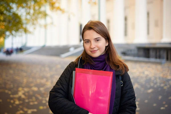 Beautiful smiling junior female student going to the classes in the cool morning of October. Portrait of a lovely student near conventional university