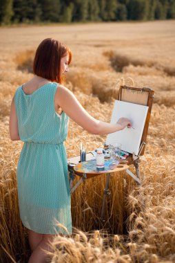 Field of ripe wheat on sunset time, girl artist drawing a picture. Warm summer time, female painter makes a piece of art, using oil paints.