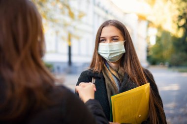 Two friends students meet each other during pandemic, greet with fists in front of university building. Students life during pandemic