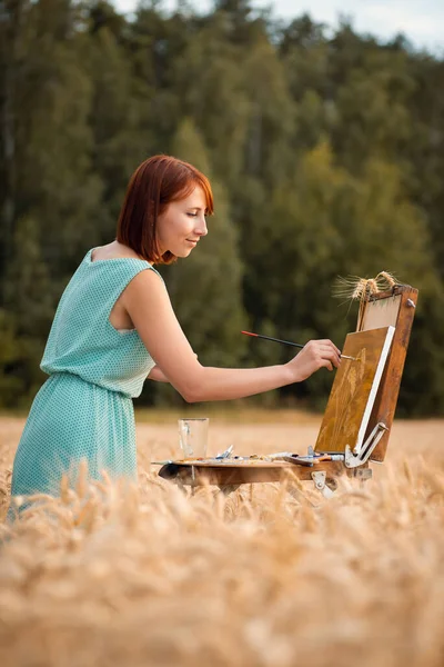 Professional painter girl working on a painting in the field of ripe wheat. Calm and joyful artist finishing drawing, vertical photo.