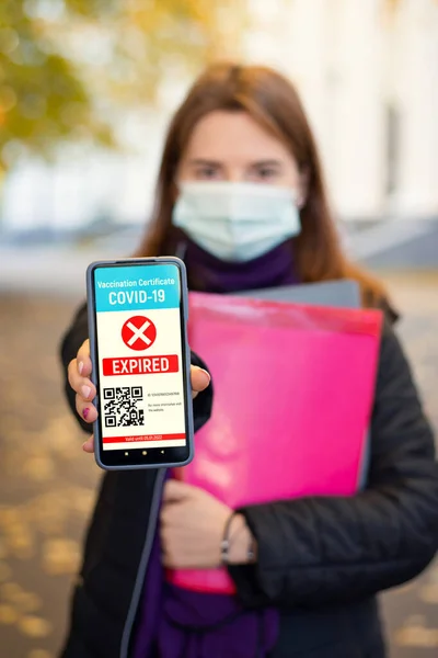Student girl with expired vaccination certificate. Young girl in medical mask standing near university building showing to the camera red expired electronic certificate