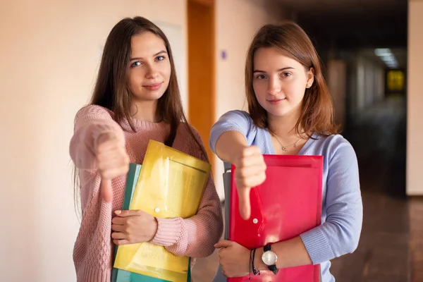 Portrait of two unhappy university students indoor in the university building showing thumbs down, unsatisfied with studying in the university. Unhappy higher education concept