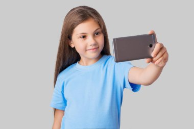 Küçük bir kızın portresi. Gri arka planda yeni bir akıllı telefonla selfie çekiyor. Mavi tişörtlü küçük kız kendi fotoğrafını çekiyor.