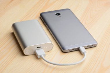 Modern gümüş akıllı telefon elektrik bankasıyla şarj edilirken ekranı açık ahşap bir masaya uzanır.