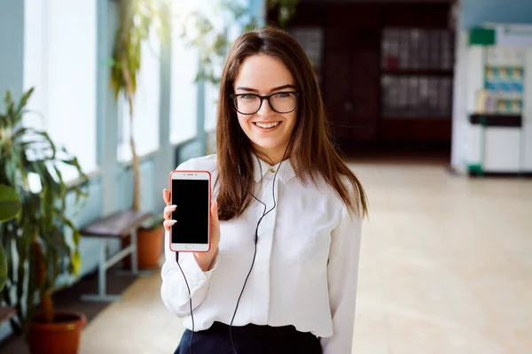 Beautiful smiling girl with smartphone displays screen of phone to the camera wearing spectacles white blouse and black skirt standing inside the university campus