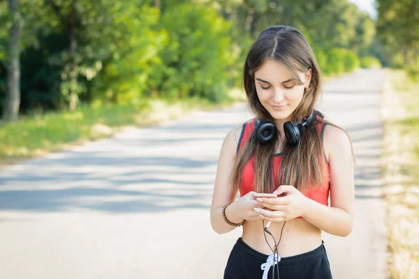 Young female using mobile phone and headphones while having a walk in the countryside in the evening. Girl wearing red t-shirt and black shorts goes along the road