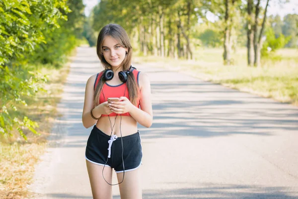 黒いショートパンツを着た幸せな10代の女の子とスマートフォンを持っている赤いTシャツ カメラを見て肩にヘッドフォンを身に着けています 田舎の夜の美しい女性 — ストック写真