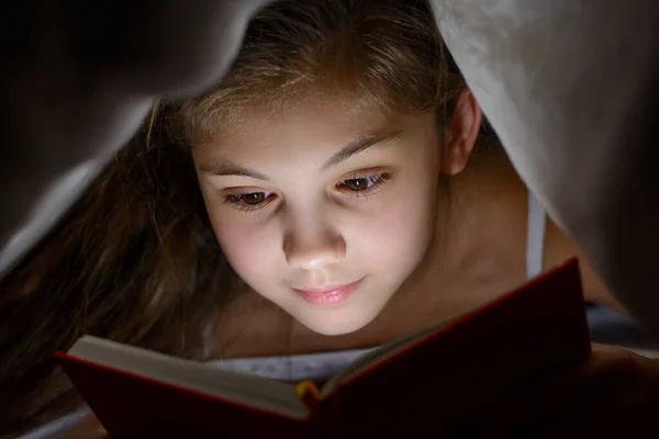 Small girl reads book with a flashlight under the blanket in bed. Beautiful caucasian child is eager to read interesting stories before bedtime