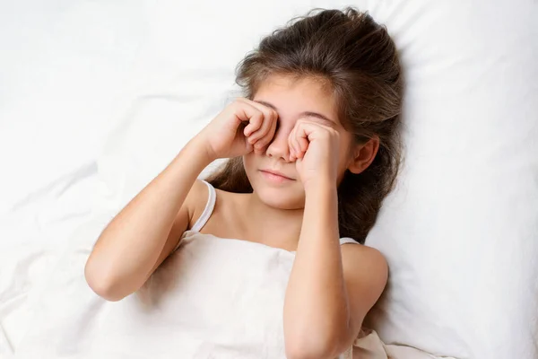 睡梦中的漂亮小女孩早上揉揉她的眼睛 有一头长长的黑发的可爱的小女孩躺在舒适的床上 揉揉着眼睛 打呵欠 — 图库照片