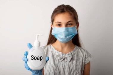 Tıp maskeli kız öğrenci ve eldivenler stüdyoda sıvı sabun şişesini gösteriyor. Grip salgını sırasında ellerini temiz tutma kavramı