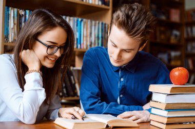 Kütüphanede iki arkadaş, masada oturuyorlar ve bilgi arıyorlar. Kız gülümsüyor ve kitaptaki bir şeye işaret ediyor, oğlan konsantre olmuş görünüyor. Eğitim ve arkadaşlık kavramı.