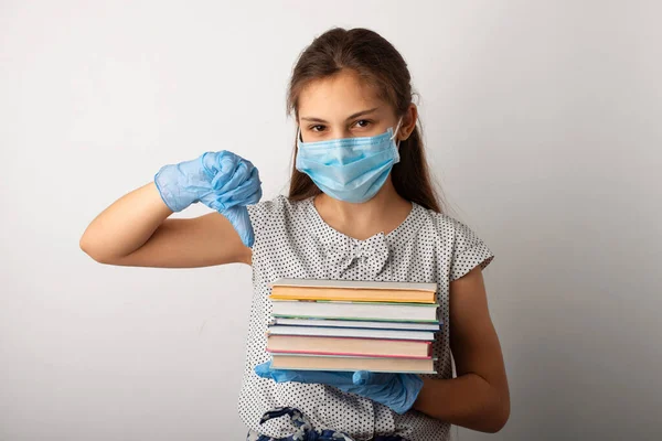 Small girl in medical mask and gloves standing with stock of books, showing thumbs down. Bored learner doesn't want to study at home during the quarantine