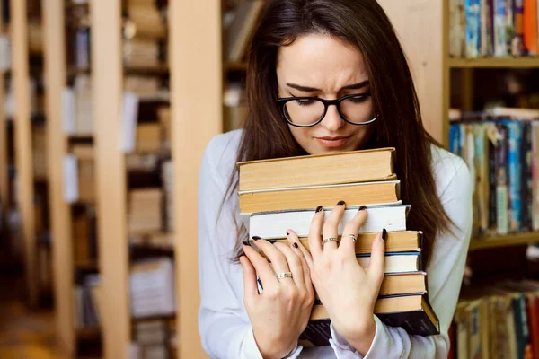 Sad female brunette student in glasses holds pile of books, looks sad and tired of learning.