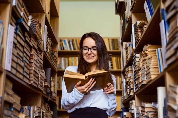 美しいブルネット学生女の子スタンドにザ図書館の間に棚の本でいっぱいと何かを読みます彼女の顔に笑顔 — ストック写真
