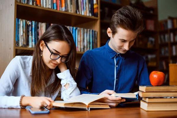 Concentrated male and female students during learning process at the library. Boy and girl sit at the table full of learning materials and read books.