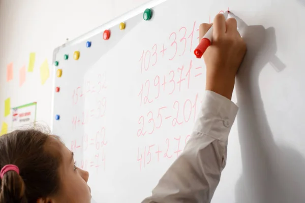 Small schoolgirl in a white blouse with pigtails writes with red marker on white board. Pupil solves mathematical equations. Colorful magnets on the board.