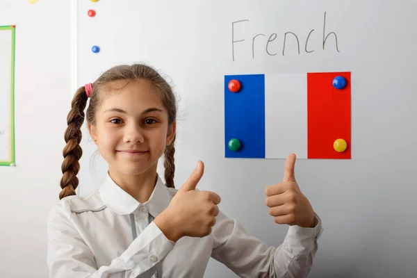 Cheerful young pupil of secondary school in white uniform with pigtails standing in front of white board and showing thumbs up. The flag of France on the blackboard. Concept of learning languages.