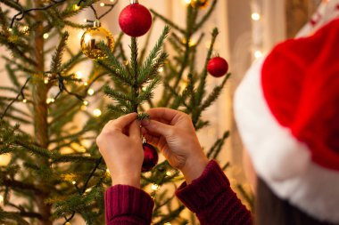 Noel ağacına büyük kırmızı bir mücevher asarken bir kızın elleri. Kırmızı Noel Baba şapkalı bir kızın arka görüntüsü.