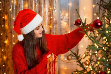 Kırmızı kazaklı ve noel şapkalı neşeli kız, sarı boncuklu köknar ağacı süslemesi. Odada parlak bir çelenk ışığı, ılık bir festival havası..