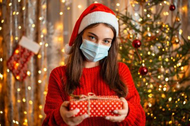 Tıbbi maskeli ve Noel Baba şapkalı genç bir kızın portresi Noel ağacının önünde duruyor ve elinde güzel dekore edilmiş bir hediye kutusu var. Kış tatilleri salgın sırasında, yeni yıl tecritte.