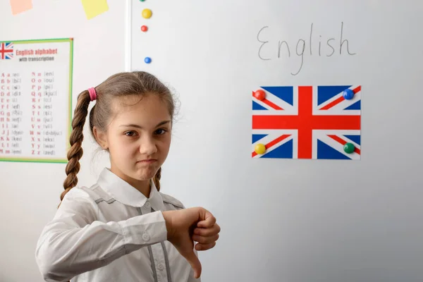 Education, learning english, no fun. Girl stands near the flag of Great Britain and shows thumb down, has dissatisfied facial expression.