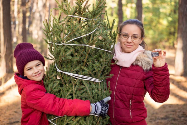 Cheerful siblings hugging a christmas tree, girl showing a chip for the tree, as a provement of legal shopping. Happy family outdoor on a bright winter day before holidays.