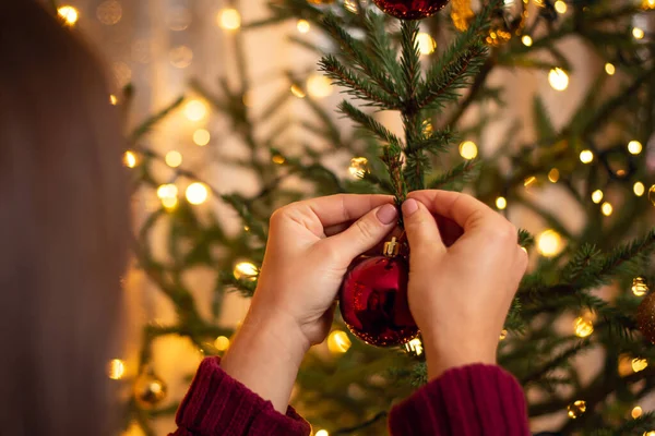 Back view of a brunette girl in sweater of burgundy colour hanging a red ball on the christmas tree. Holidays, festive mood concept.