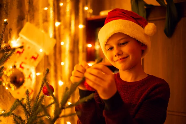 Smiling boy in christmas hat decorating the fir beauty in the room, full of yellow lights from garlands. Warm festive mood, holidays spirit.