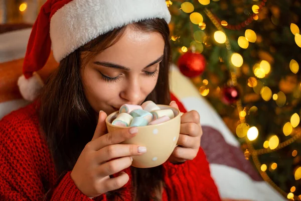 Beautiful girl in red Christmas hat drinking tasty cocoa with marshmallow on top near decorated christmas tree. Cheerful girl enjoying comfortable Christmas atmosphere at home