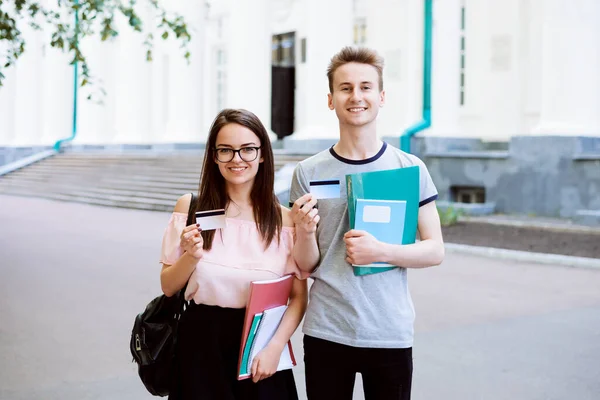 Student couple standing outdoors, smiling, looking to the camera and showing credit card. Girl and boy happy to become scholarship and being financial independent.