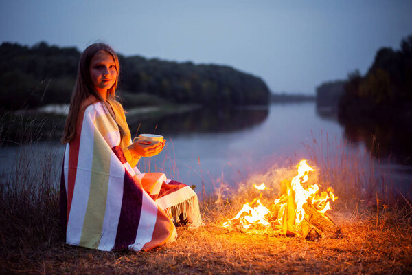 Teenage girl sitting near the bonfire with a cup of warm tea near the river in the evening. Concept of having a picnic, spending time in nature