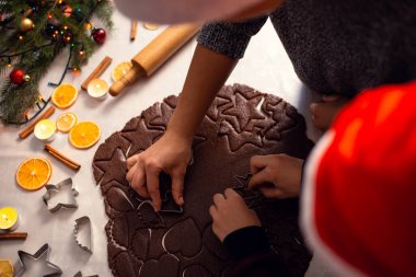 Evde Noel bisküvisi pişiriyorum. Anne ve oğlu kırmızı Noel Baba şapkalı fırına çiğ bisküvi koymadan önce siyah çikolata hamuru kesiyorlar.