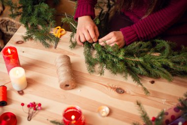 Doğal köknar ağacı dallarından Noel çelengi yapan bir kadının elleri. Mumlar, sicim, masadaki çelenkler için yapay böğürtlen..