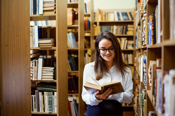 Female student with books in her hands between shelves full of books. Smiling girl likes studying and eager to enlarge her knowledge and skills