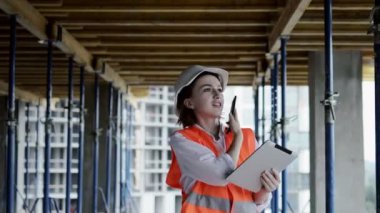 Güvenlik ceketli ve kasklı genç bayan inşaat alanında dizüstü bilgisayarla çalışırken cep telefonuyla konuşuyor..