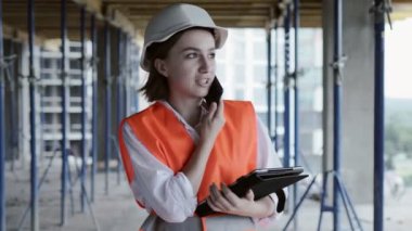 Güvenlik ceketli ve kasklı genç bayan inşaat alanında dizüstü bilgisayarla çalışırken cep telefonuyla konuşuyor..