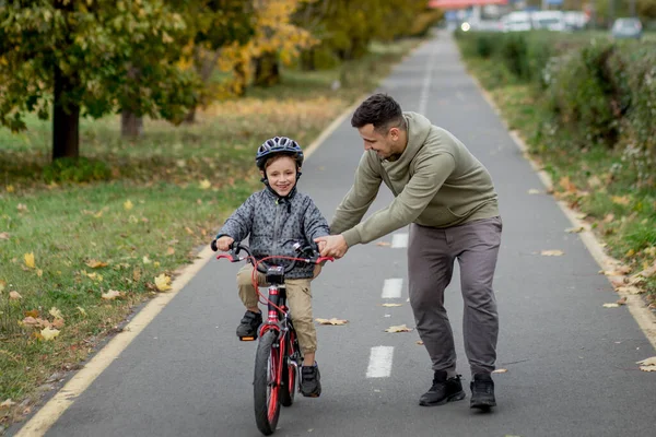 Pai ensina seu filho a andar de bicicleta no caminho da bicicleta no parque. O pai está segurando uma bicicleta e o filho está sentado nela. — Fotografia de Stock