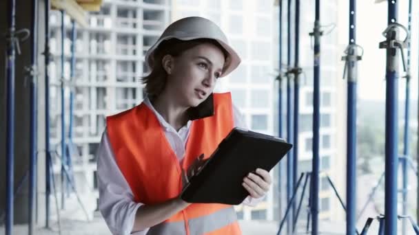 Young female civil engineer in safety jacket and helmet is talking on mobile phone on development construction site outdoor while working on laptop — Stock Video