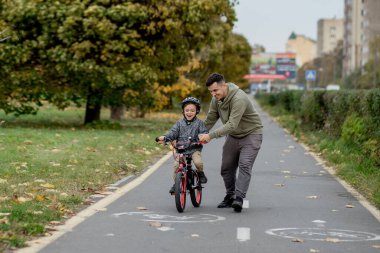 Baba oğluna parktaki bisiklet yolunda bisiklet sürmeyi öğretiyor. Baba bir bisiklet tutuyor ve oğlu üzerinde oturuyor..