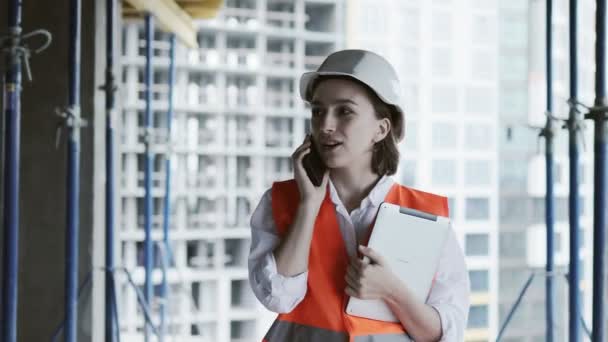 Young female civil engineer in safety jacket and helmet is talking on mobile phone on development construction site outdoor while working on laptop — Stock Video